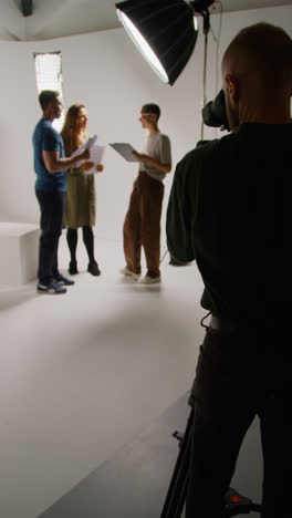 Vertical-Video-Of-Film-Camera-Operator-Shooting-Director-Talking-With-Male-And-Female-Actors-Holding-Scripts-Rehearsing-For-Shooting-Movie-Or-Video-In-Studio-1